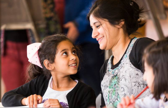 One woman looks down at a child who is doing art work at a table. They both have black hair and brown eyes, there are people in the background which are blurry.