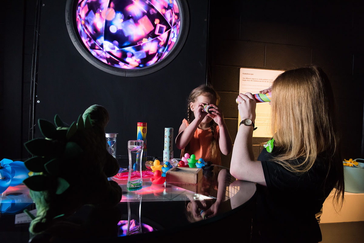 Children sit at a table looking through cardboard periscopes. In the background there is a multicoloured light behind them. On the table they sit at, there are lots of craft materials.