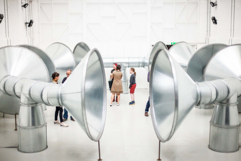 The image shows a large warehouse space with huge silver tubes. At the end of each tube is a large open bowl, which looks like the end of a trumpet. There are figures in the middle of these tubes.