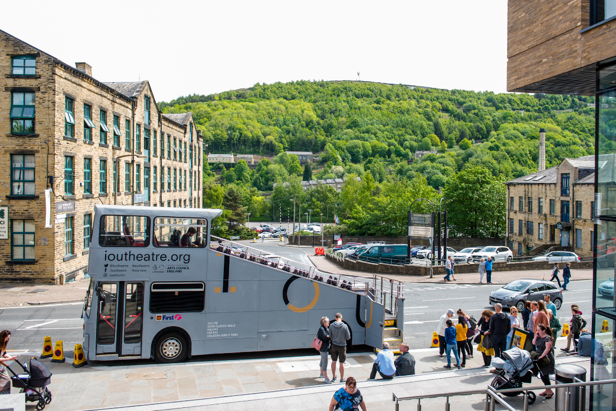 A grey bus with a diagonal section cut of out it, to make it open top is parked infront of a large mill. There are trees in the distance, and a queue of people waiting to board the bus. It's a sunny day.