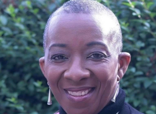 This is a headshot of a woman who is looking at the camera. She is smiling and is wearing long, sparkly earrings. Her skin is brown, and she has a shaved head and is wearing a black shirt.