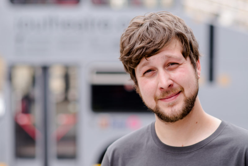 A man in his 30s is looking at the camera. He has white skin, and has brown hair which falls around his ears. He is smiliing with his mouth closed, and has a beard. In the background, there is a grey bus.