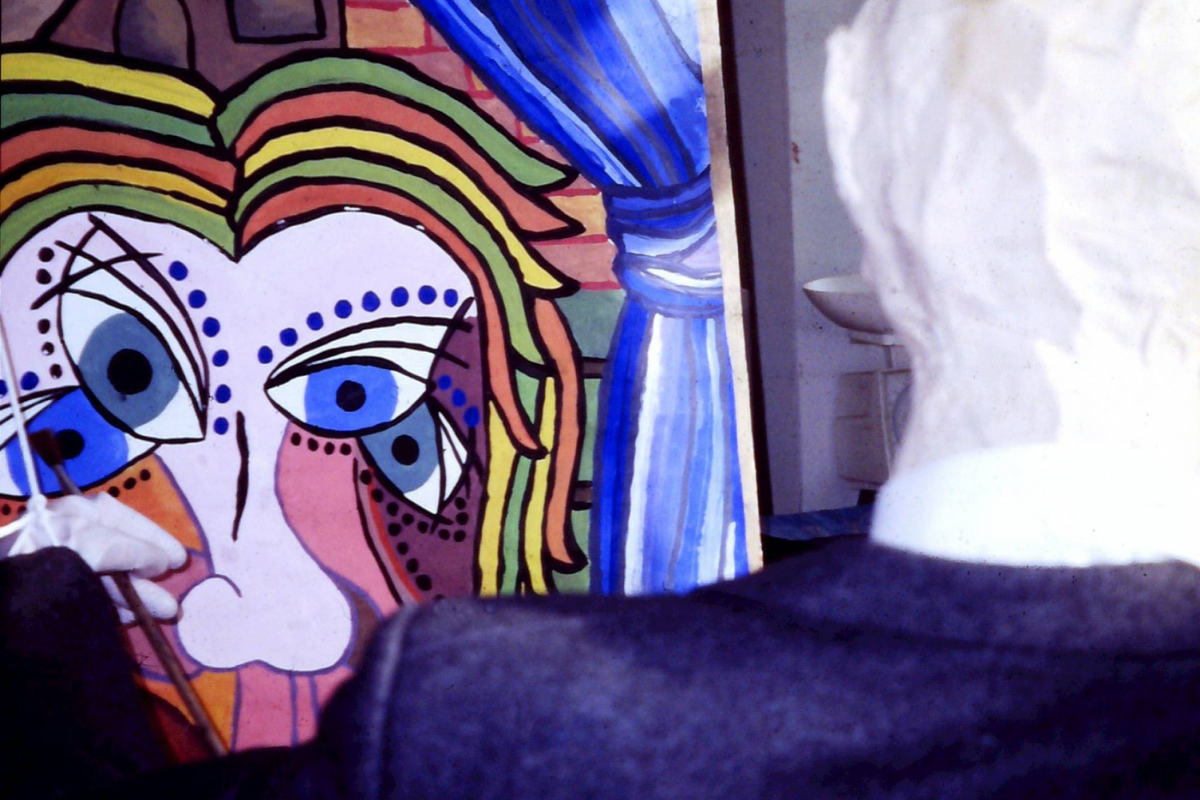 A large drawing sits in the back of a room. It's backlit and colourful and depicts a face with long black eye lashes.