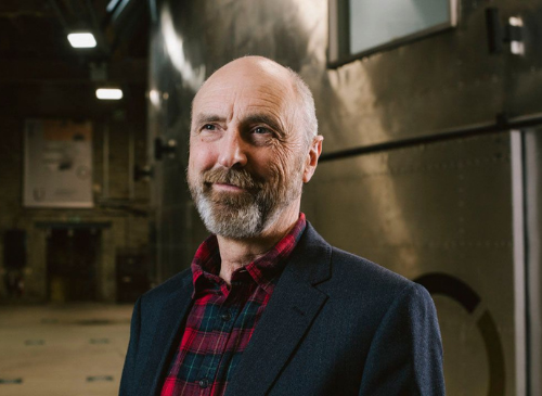 A white man stands looking just away from the camera. He has a bauld head and a grey beard. He is smiling and wears a navy jacket with red and black checked shirt underneath. In the background there is a metal and brick wall.