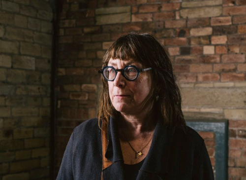 A woman with glasses has brown hair and is looking away from the camera. There is a brick wall behind her. She is white and wears a gold necklace.