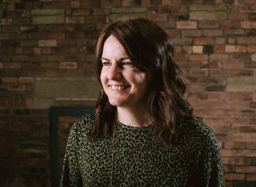 A woman looks to the left of the screen with a brick wall in the background