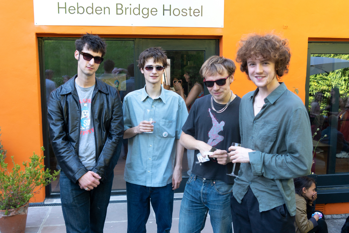 Four young men stand in front of an orange wall. There is a sign above them which has the words 'Hebden Bridge Hostel' written on a sign. They all have sunglasses on, apart from the one on the far right who isn't wearing sunglasses, he is holding a glass of white wine in his hand.