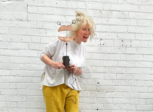 There is a woman smiling, with blond hair, she is holding a metal sculpture that is shaped like lips, it is made from copper coloured metal and has two metal eyes at the top.