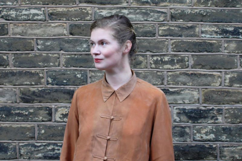 A woman stands in front of a brick wall. She is wearing a rust coloured shirt and looking to the left. She has white skin and blonde hair which is tied back.