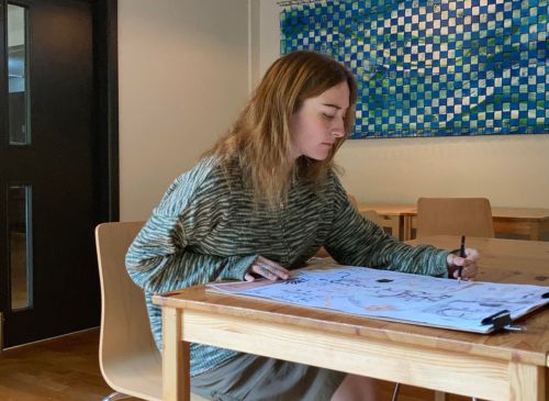 woman doing artwork at wooden desk she has long hair and wearing a green sweater